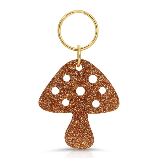A mushroom-shaped keychain with brown glitter and white spots hangs from a gold ring.
