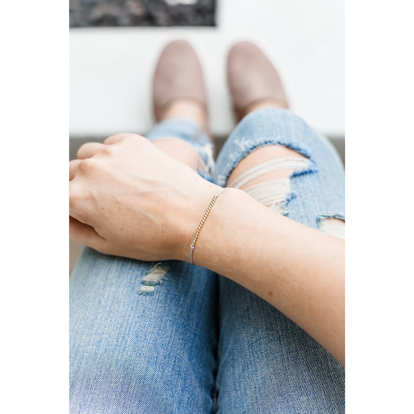 A person wearing a delicate gold bead bracelet sits with their legs crossed wearing distressed jeans and taupe slip-on shoes.