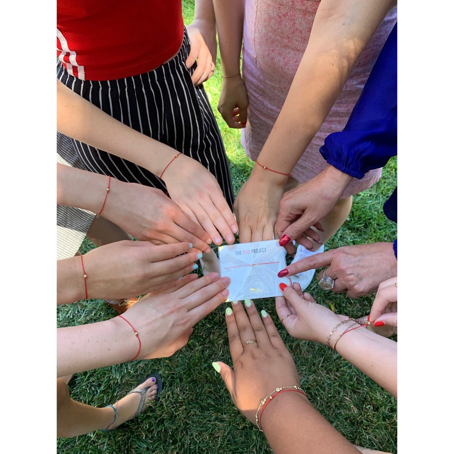 A group of people place their hands, all wearing red string bracelets, around a card for The Pad Project.