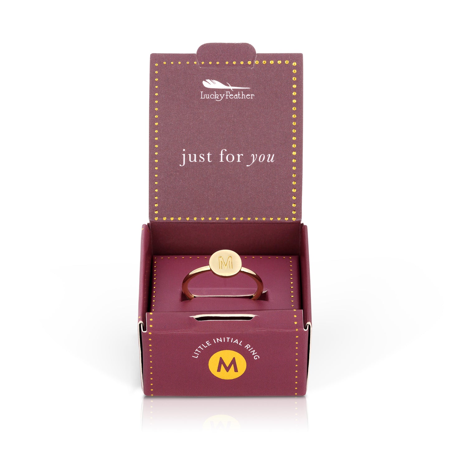 A gold-colored ring with the letter "M" is presented in a purple box with gold dots.