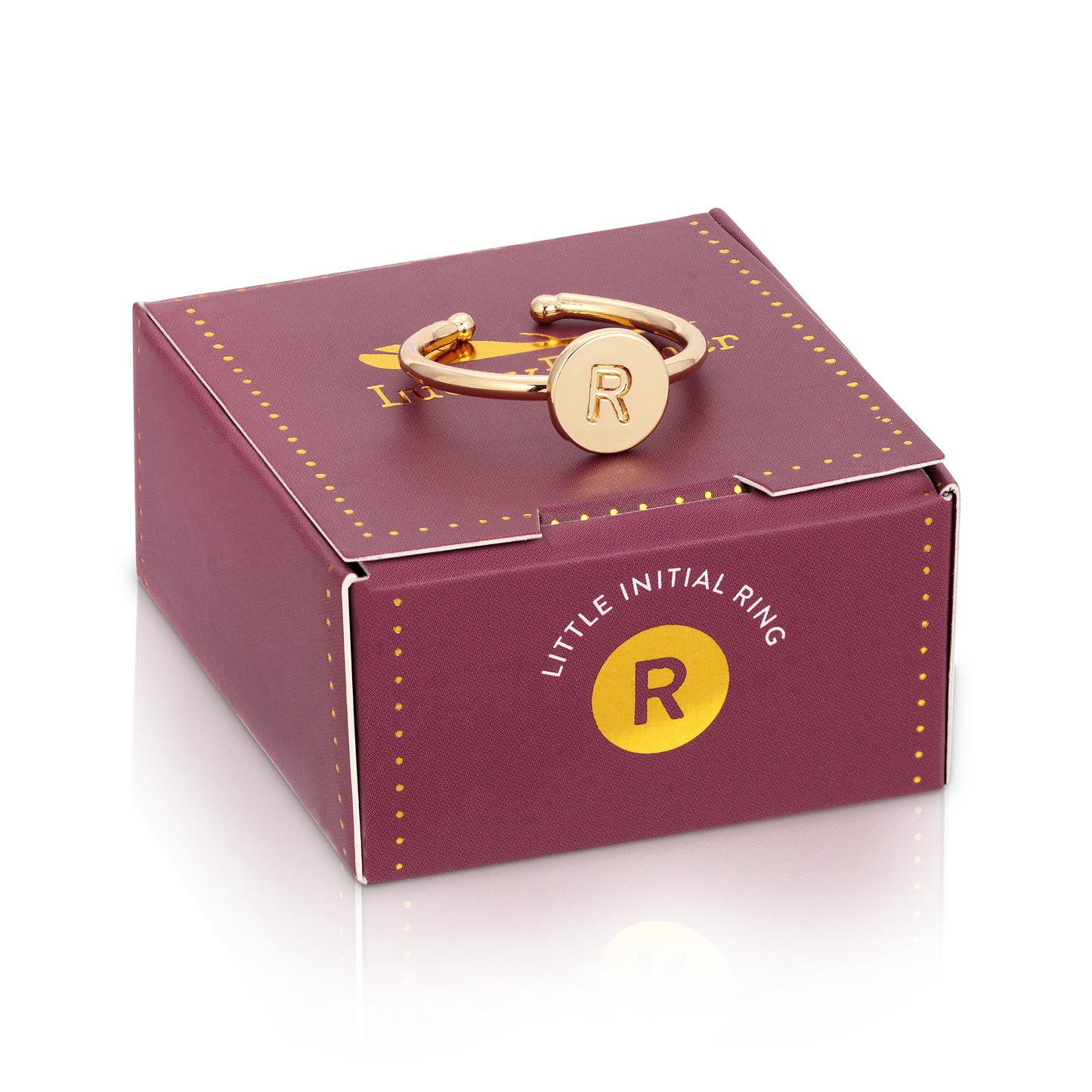 A gold-colored ring with the letter "R" on a circular pendant sits atop a purple box labeled "LITTLE INITIAL RING," also featuring a golden "R" symbol.