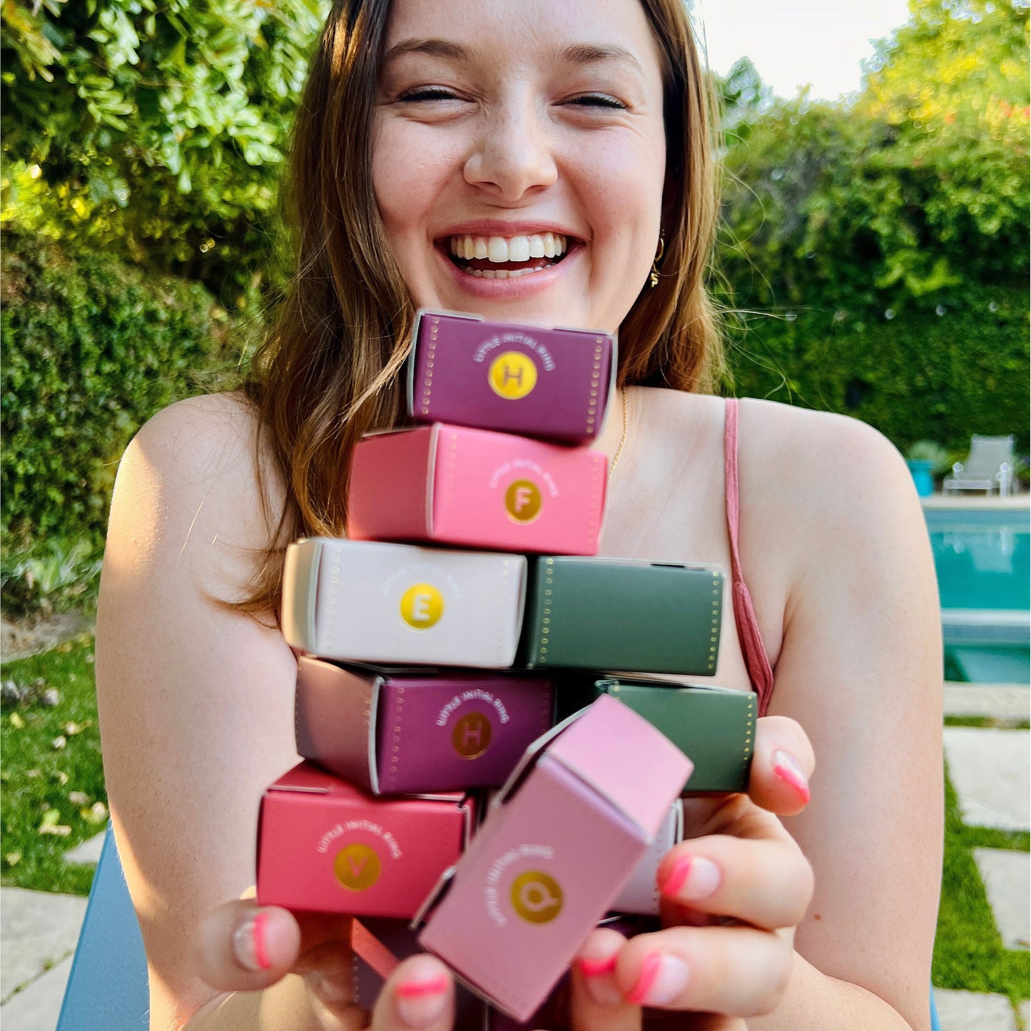 A woman smiles broadly while holding a stack of colorful boxes labeled "Little Initial Ring" outdoors near a pool.
