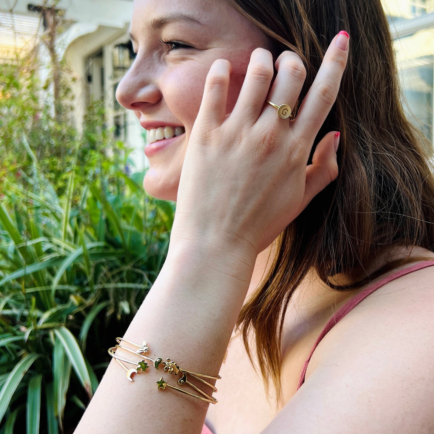 A woman with brown hair smiles and wears a gold initial ring and several whimsical gold bracelets.
