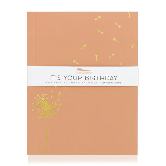An orange journal wrapped in a white band that reads "Lucky Feather IT'S YOUR BIRTHDAY MAKE A BUNCH OF WISHES AND WATCH THEM COME TRUE" is featured on a white background.