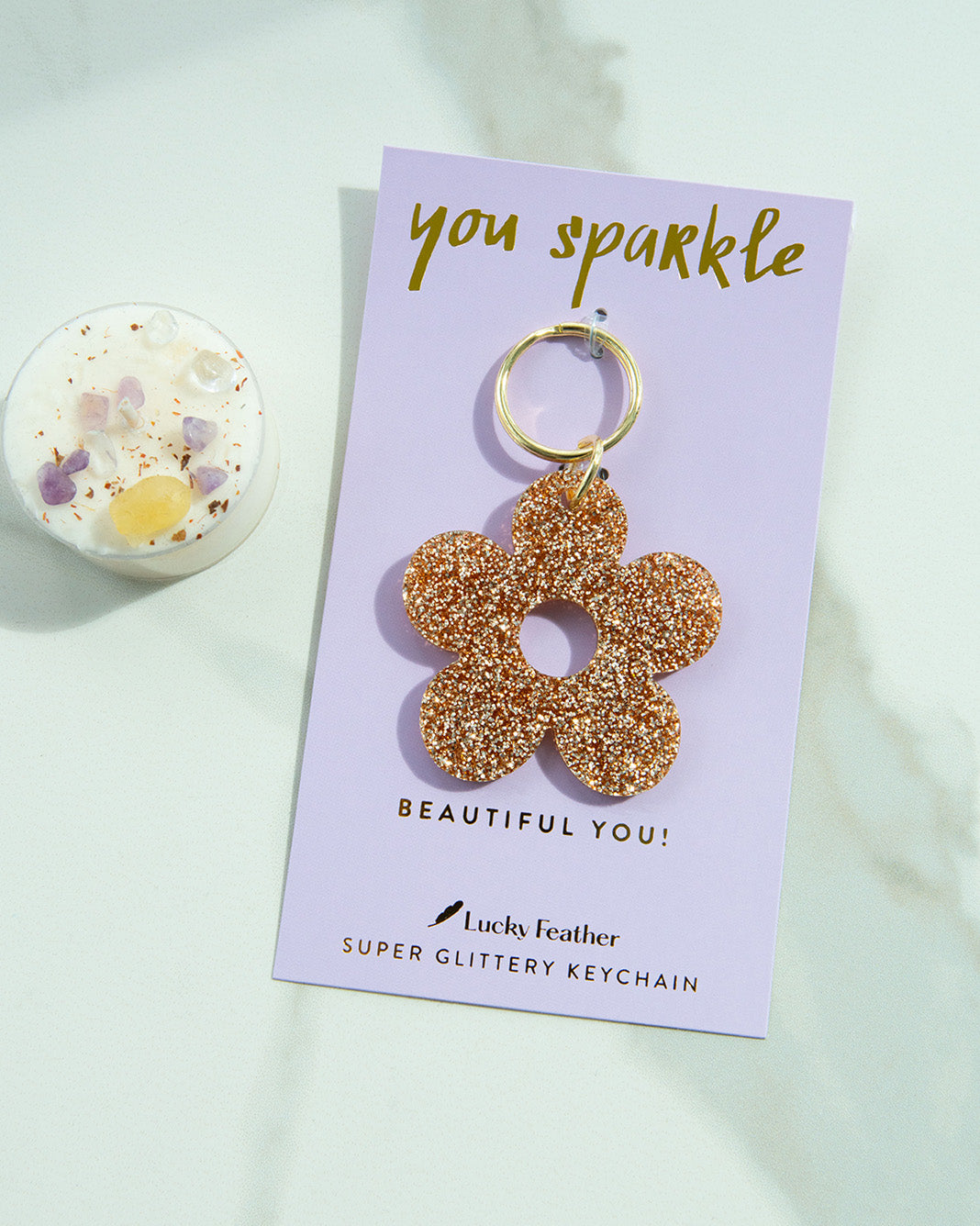 A glittery flower keychain on a lilac card that reads 'you sparkle, beautiful you' next to a small candle with crystals.
