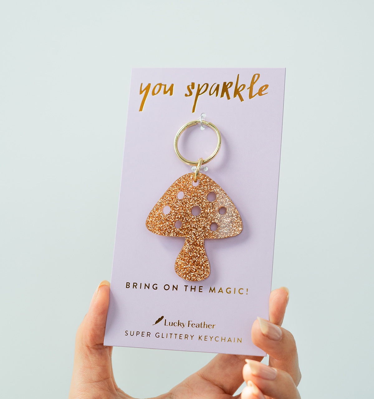 A hand holds a lavender card displaying a glittery mushroom keychain.
