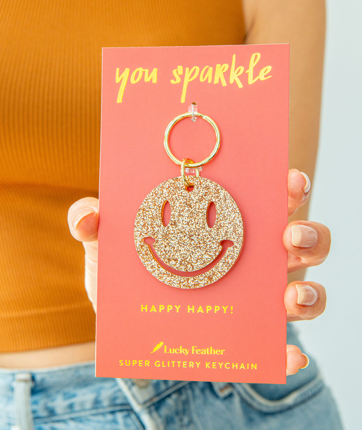 A person holds a pink card displaying a glittery gold smiley-face keychain.