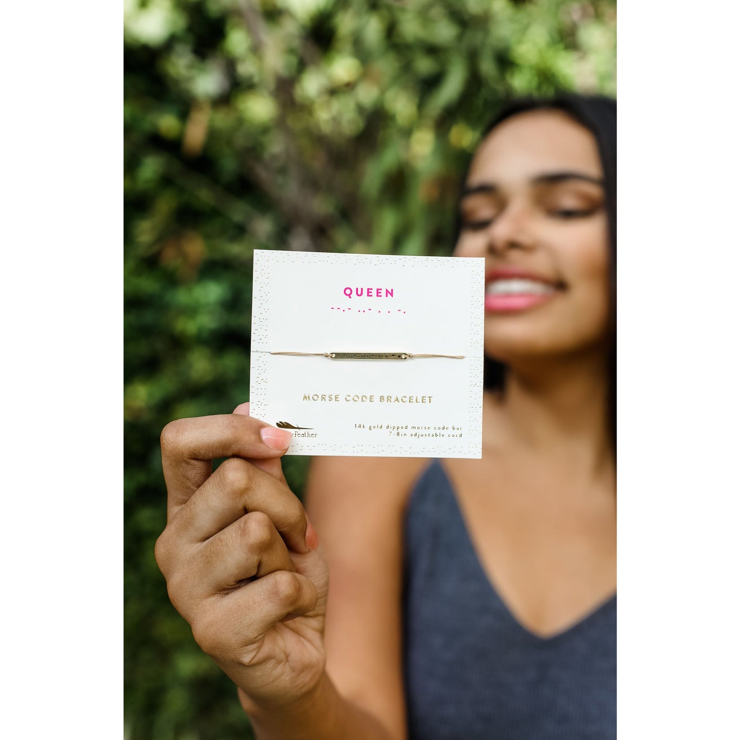 A person holds a product card for a "Queen" morse code bracelet in front of green foliage.