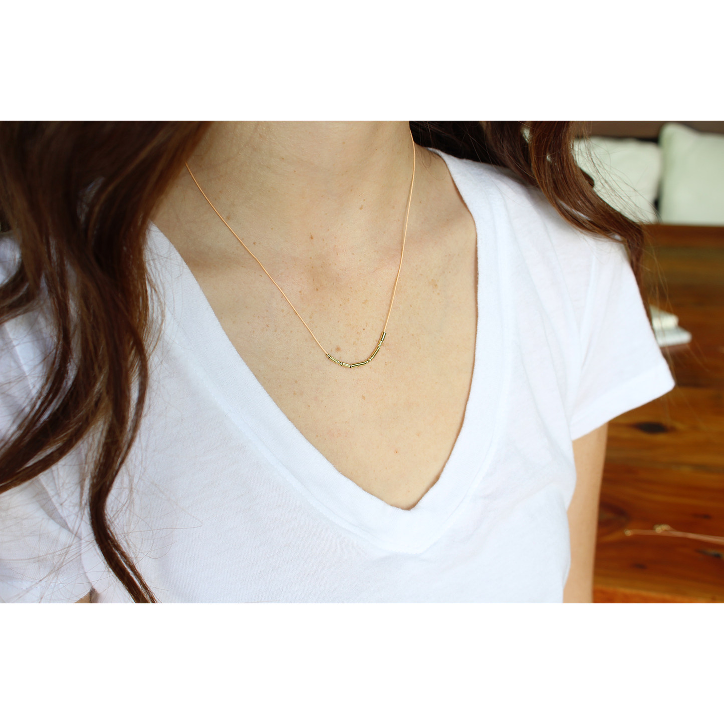 A person wearing a white v-neck shirt and a delicate gold necklace with small cylindrical beads.