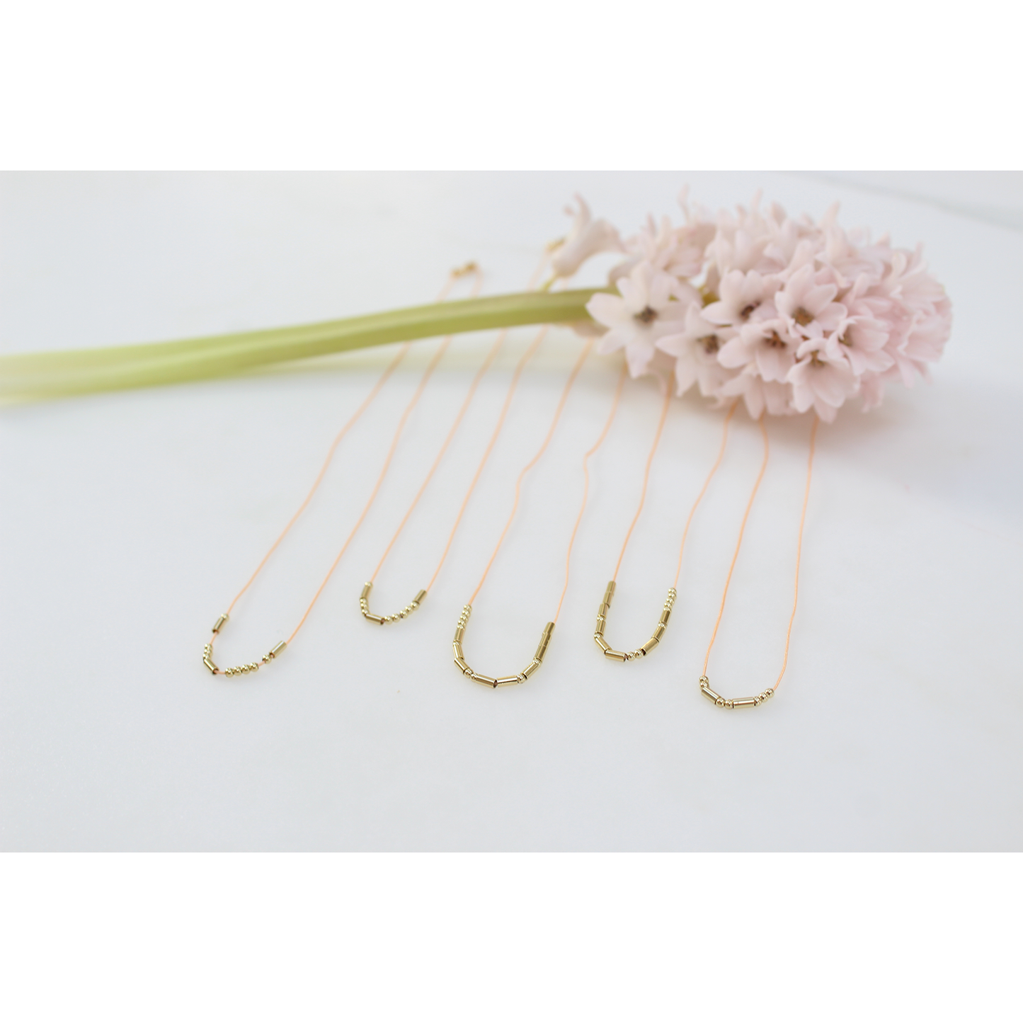 Five delicate gold necklaces are arranged next to a light pink hyacinth on a white surface.