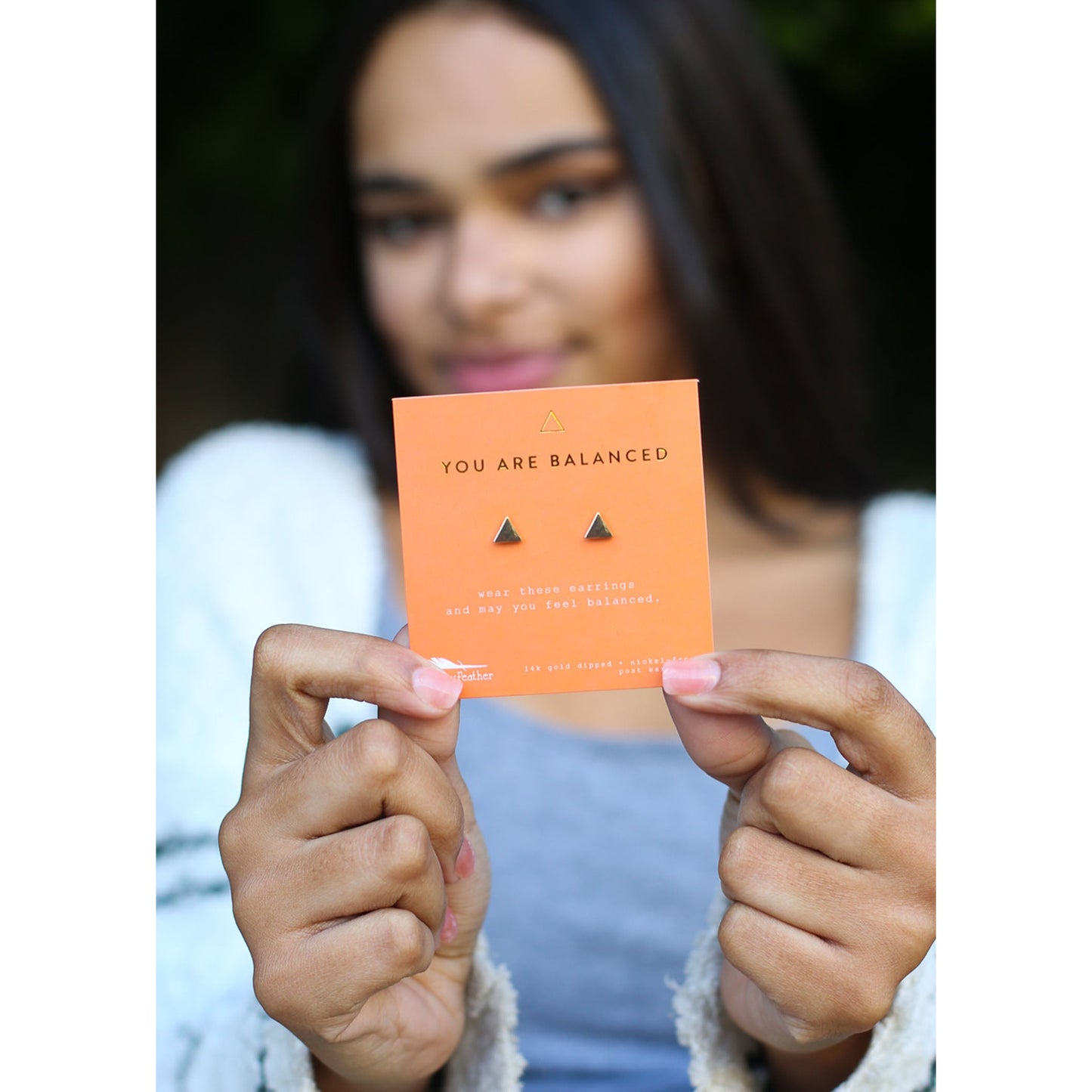 A person holds up a card of triangle earrings labeled "YOU ARE BALANCED".