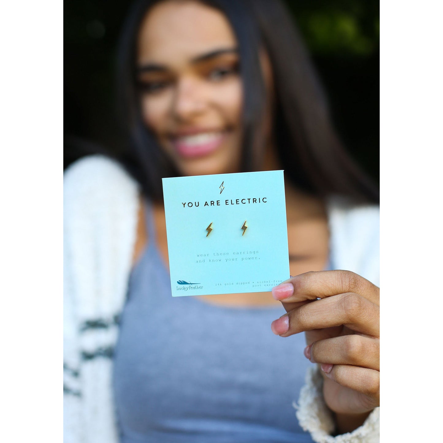 A smiling person holds a card that reads "You Are Electric" featuring small lightning bolt earrings.