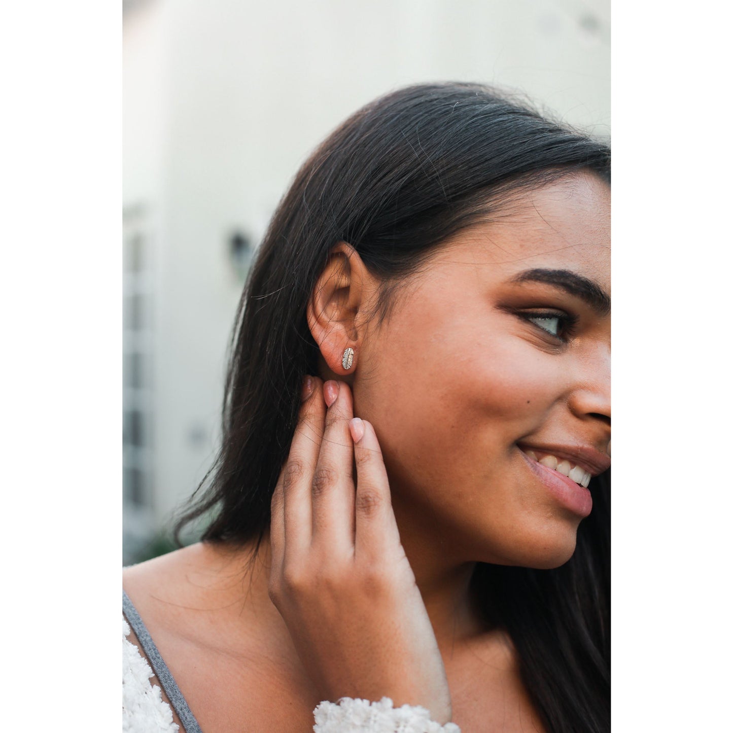 A smiling woman is touching her ear, showcasing a small feather-shaped earring.