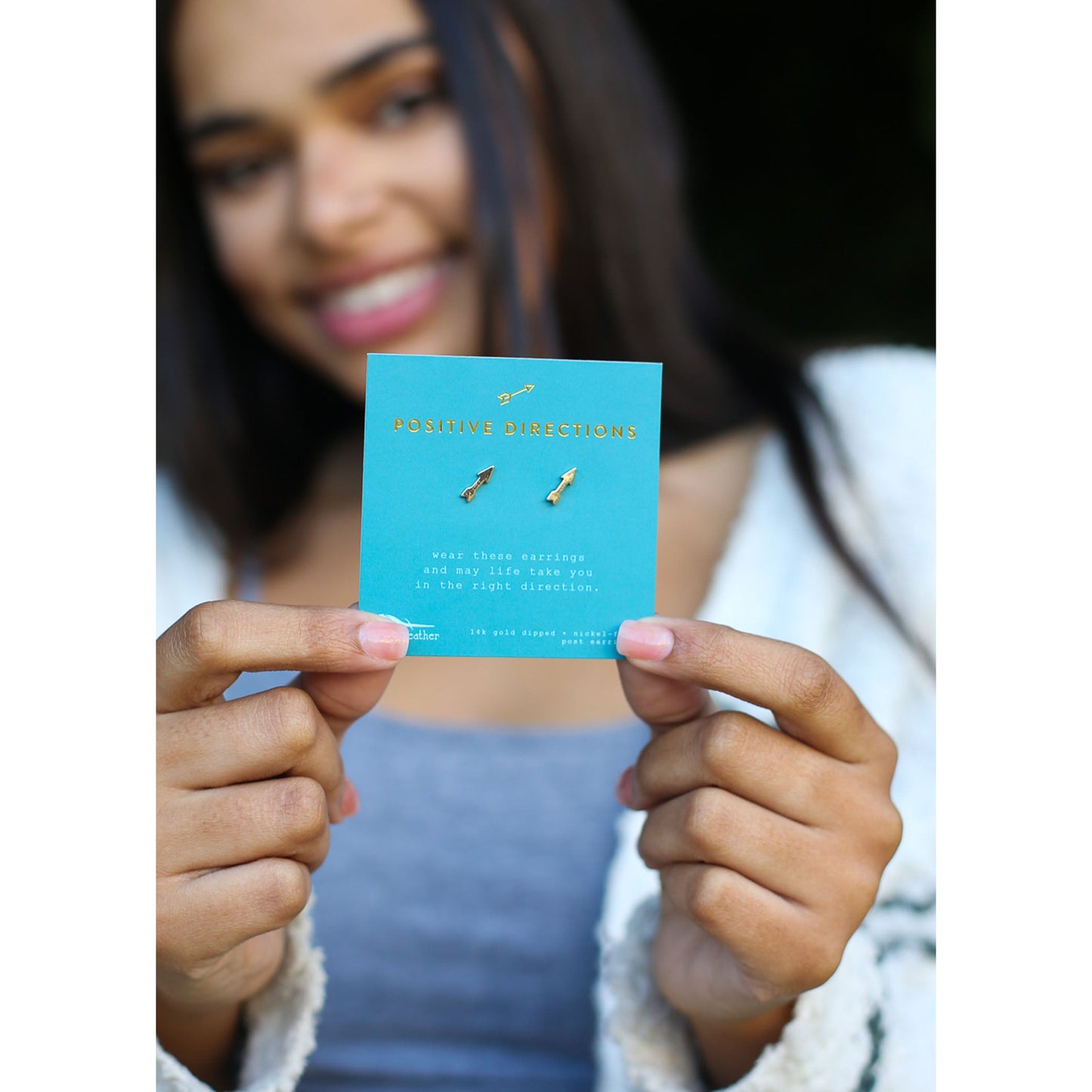 A smiling woman holds a product card for "Positive Directions" earrings, which are gold-toned arrows.