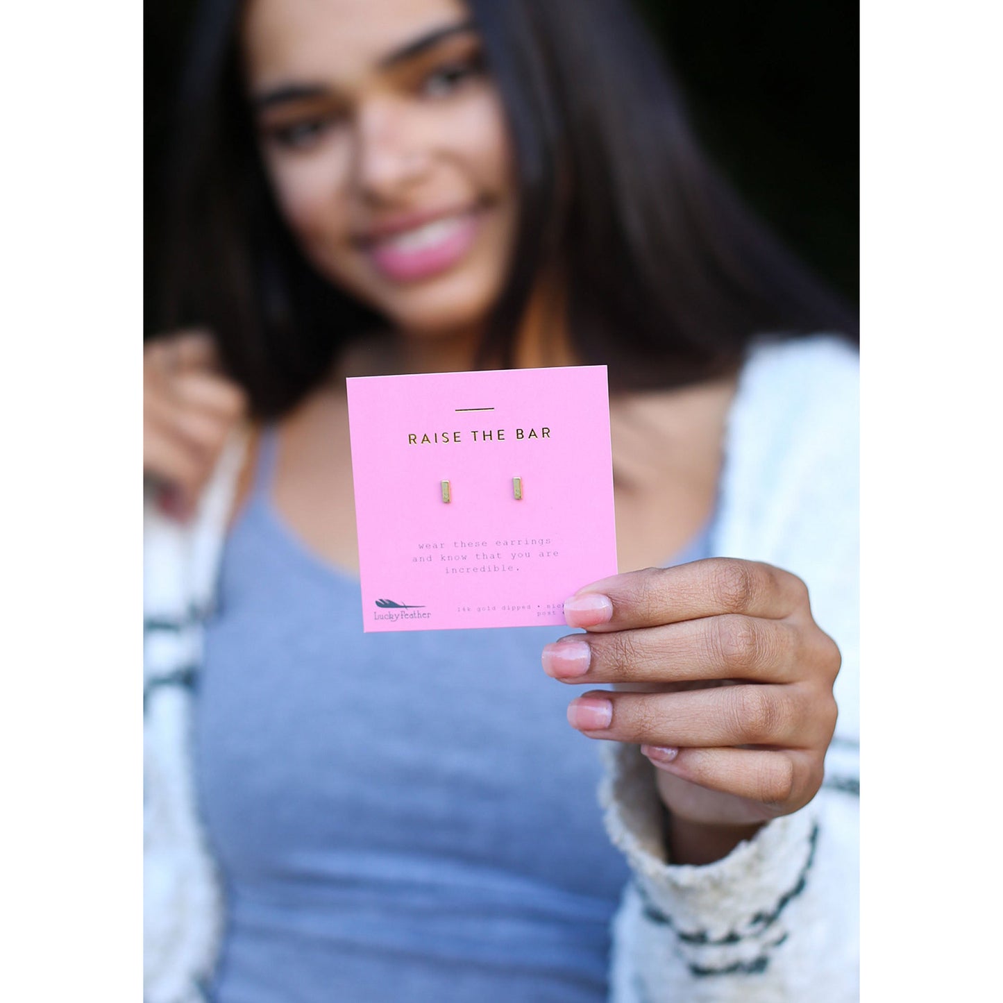 A smiling young woman holds a pink card with "RAISE THE BAR" printed on it, displaying a pair of small gold earrings.