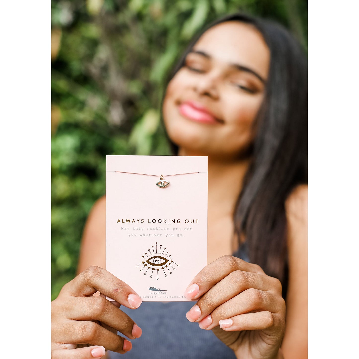 A woman holding an "Always Looking Out" necklace against an out-of-focus green background.