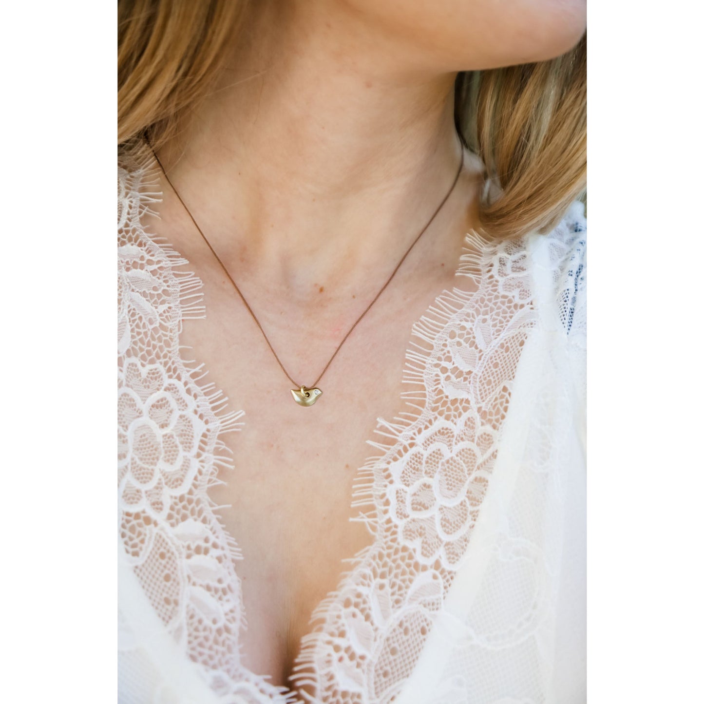 A person wearing a white lace top and a gold bird necklace with a brown cord.