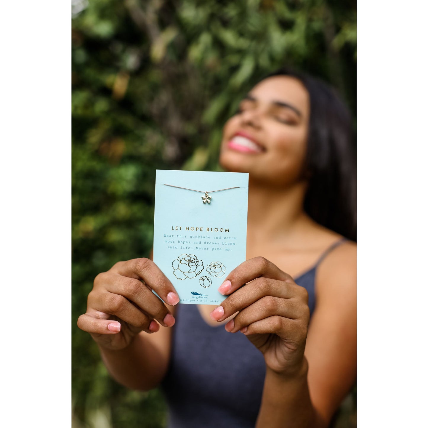 A smiling woman with closed eyes holds up a card displaying a delicate floral necklace.