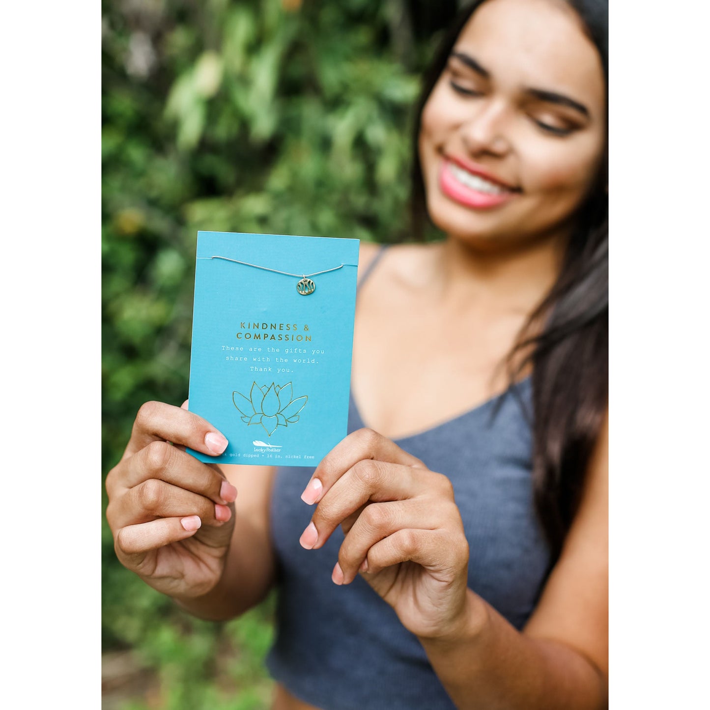 A smiling woman holds a card with a necklace that reads "Kindness & Compassion."