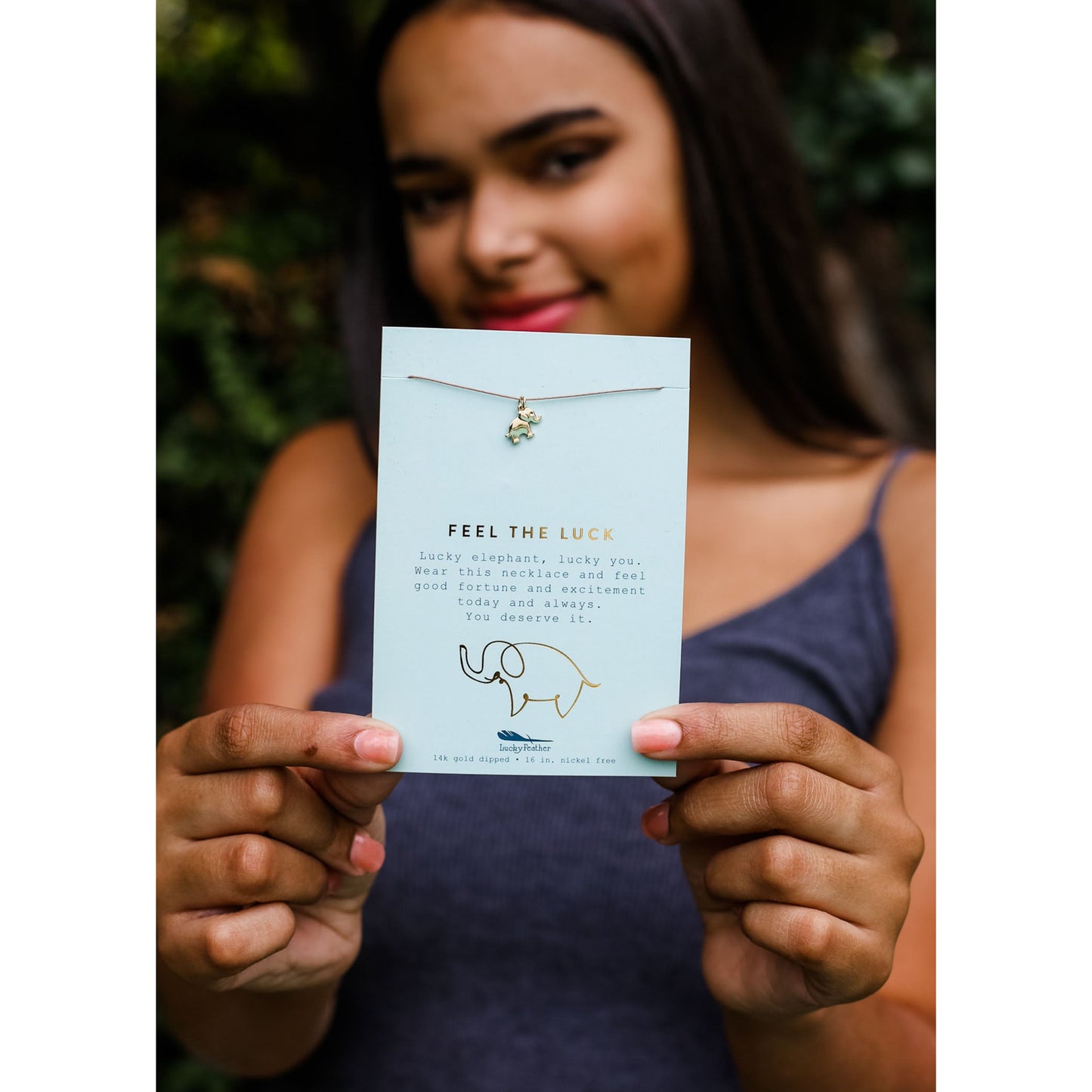 A person holds up a card with a gold elephant necklace that says "FEEL THE LUCK".