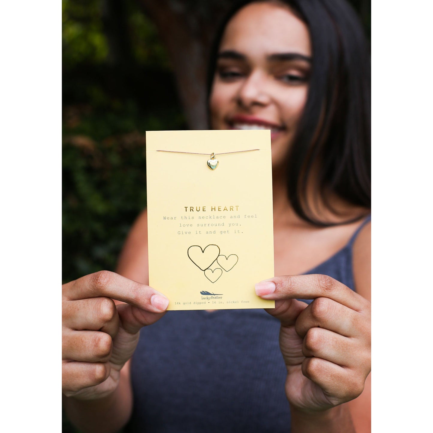 A smiling woman holds a card advertising a "True Heart" necklace.