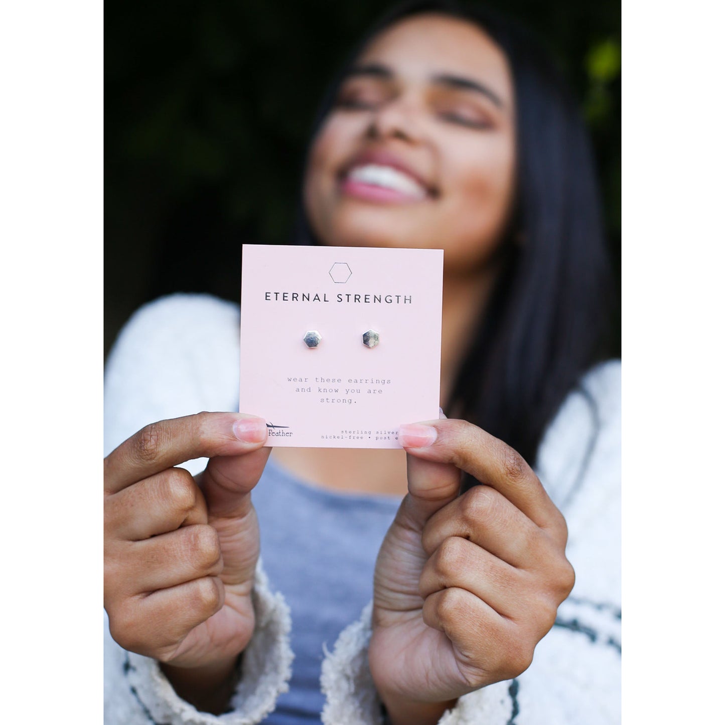 A smiling person holds a pink display card with hexagon stud earrings labeled "Eternal Strength".