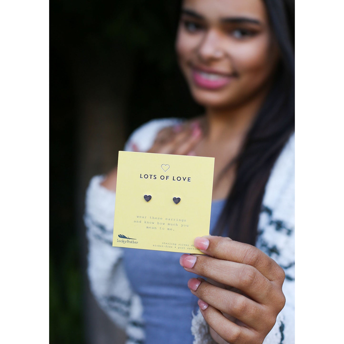 A person holds up a yellow card displaying heart-shaped earrings, with the words "LOTS OF LOVE" printed on it.