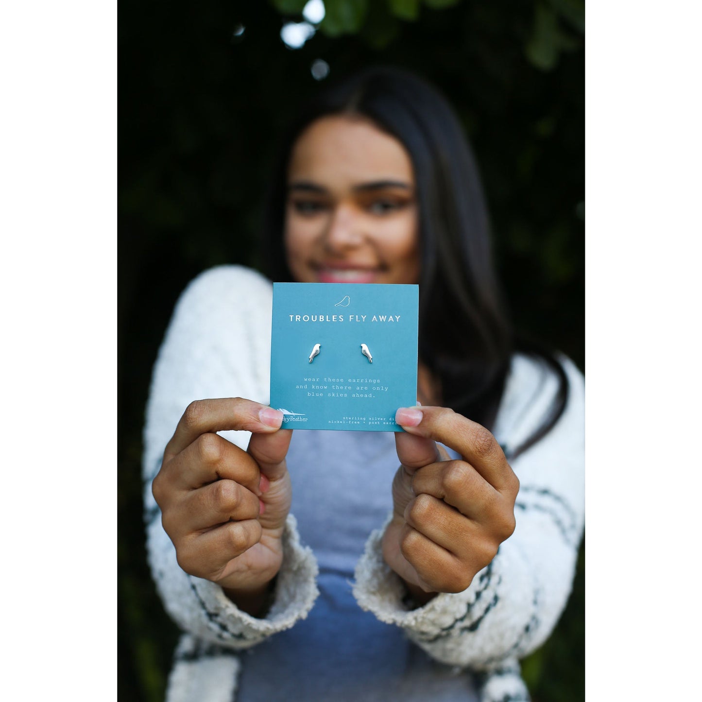 A smiling woman holds up a card displaying bird-shaped earrings with the phrase "Troubles Fly Away."
