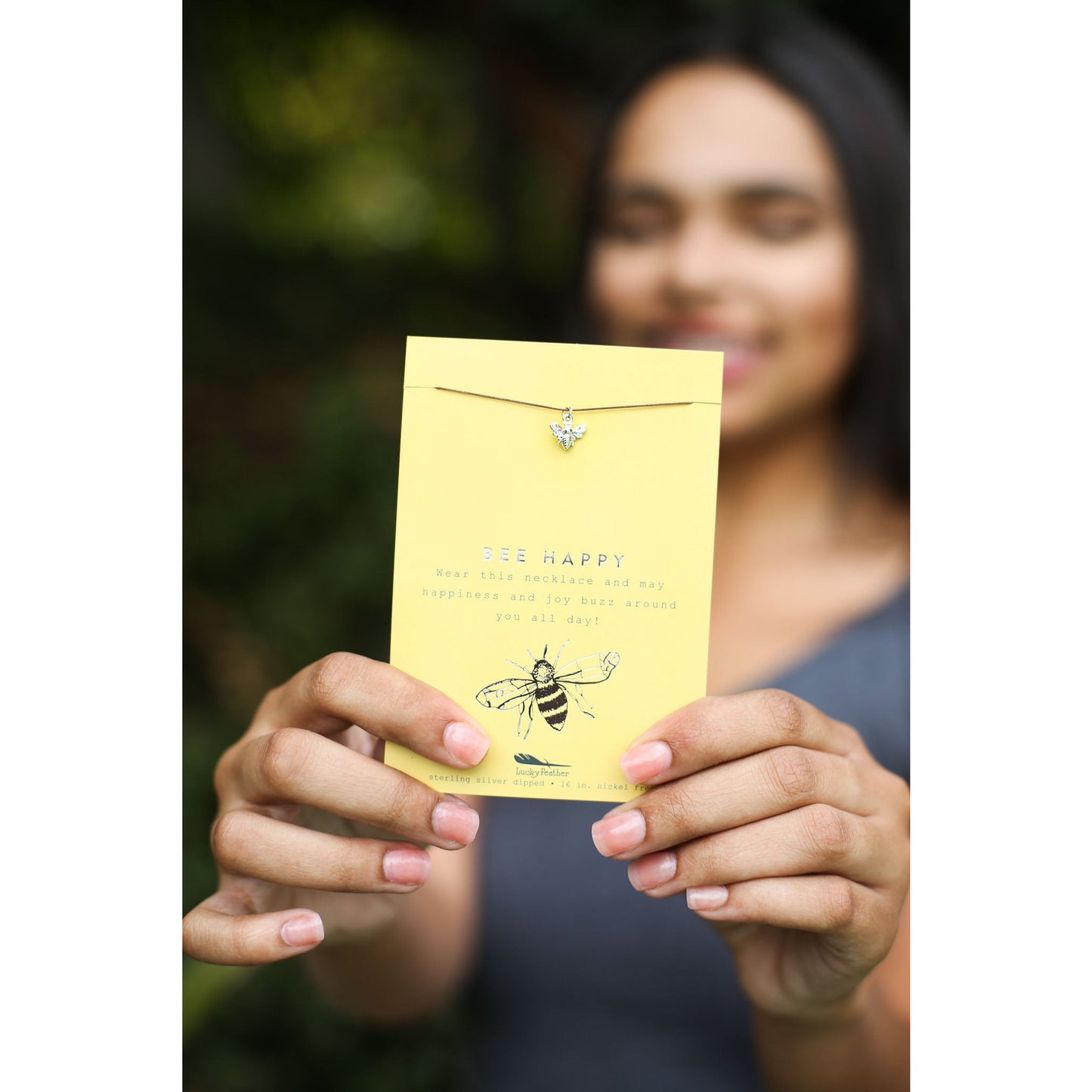 A woman holds a "Bee Happy" necklace on a yellow card.