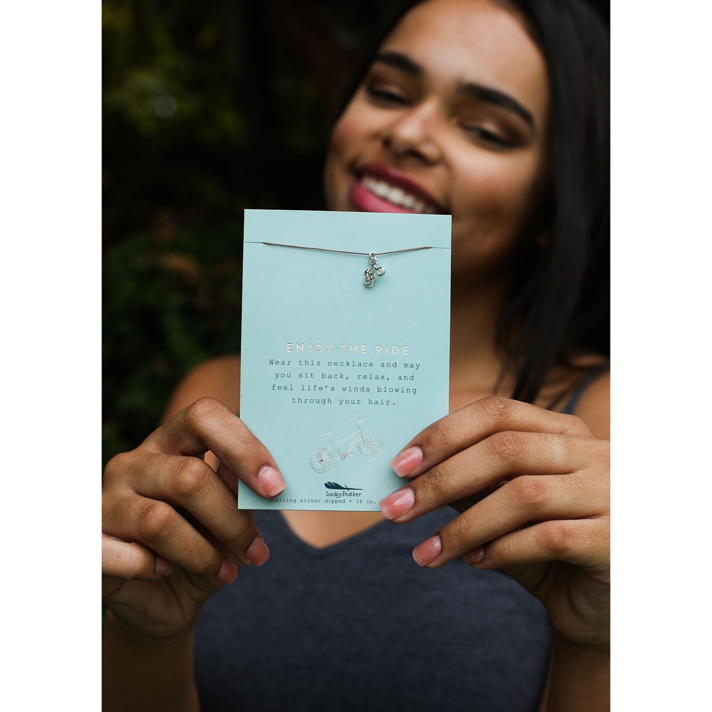A smiling person holds up a necklace on a light blue card with the words "Enjoy the ride".