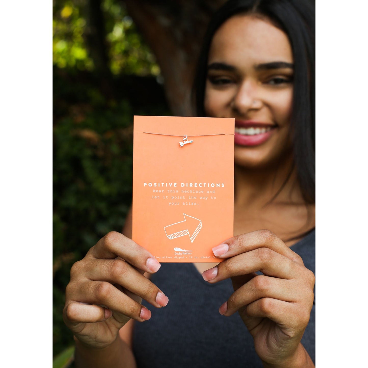 A smiling person holds up an orange card with the text "POSITIVE DIRECTIONS" and a small arrow pendant necklace.