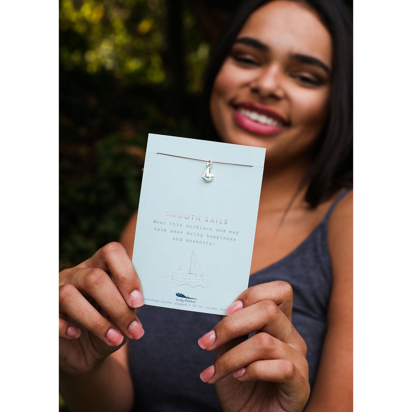 A smiling woman holds up a necklace with a sailboat charm on a card that reads "Smooth Sails."