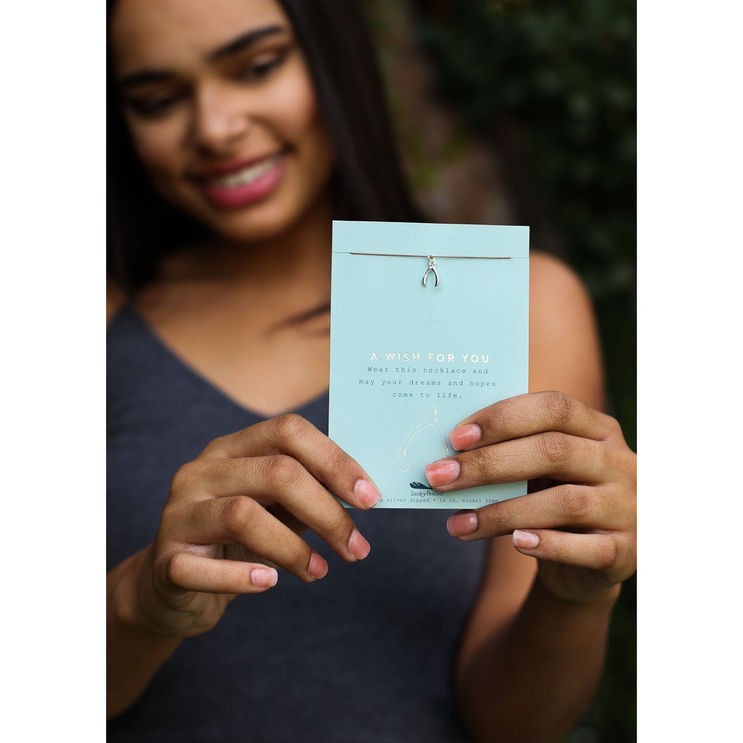 A woman is holding up a light blue card with a wishbone necklace.