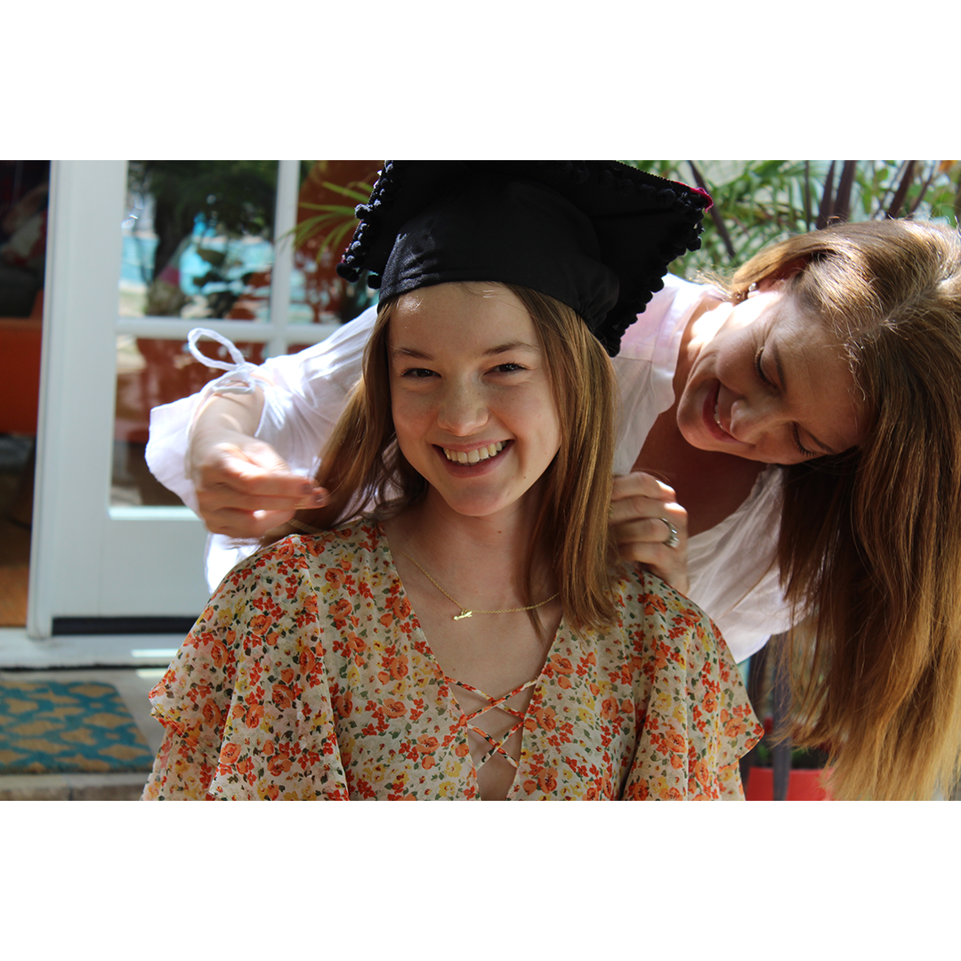 A smiling young woman in a floral top and graduation cap stands while another woman adjusts her hair.
