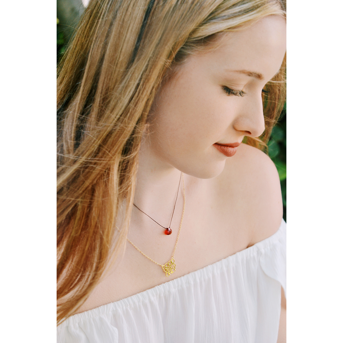 A fair-skinned woman with blonde hair, wearing a white off-the-shoulder top and two necklaces, looks down with her eyes closed.