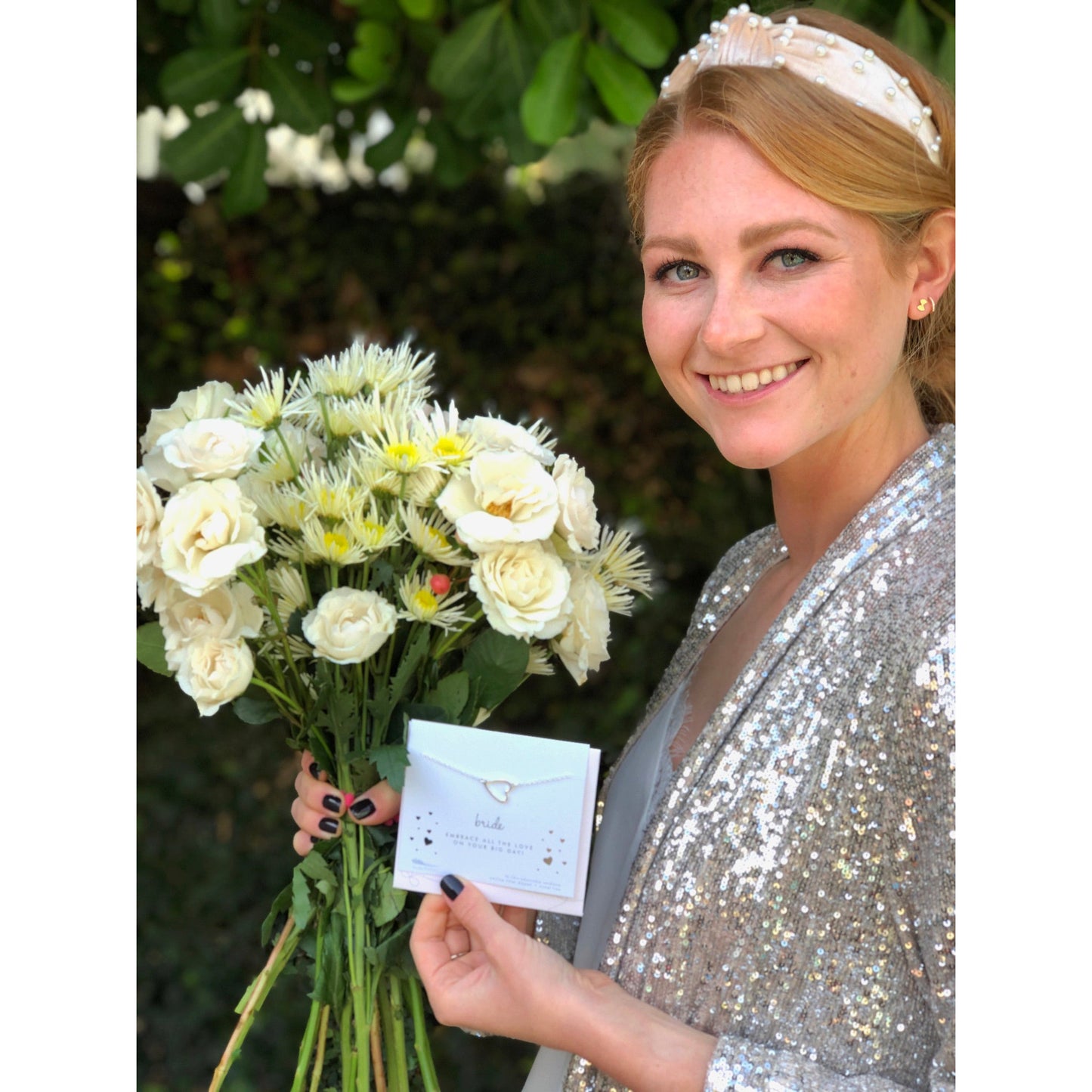 A woman with a pearl headband and sequined jacket holds a bouquet of white flowers and a card with a necklace.