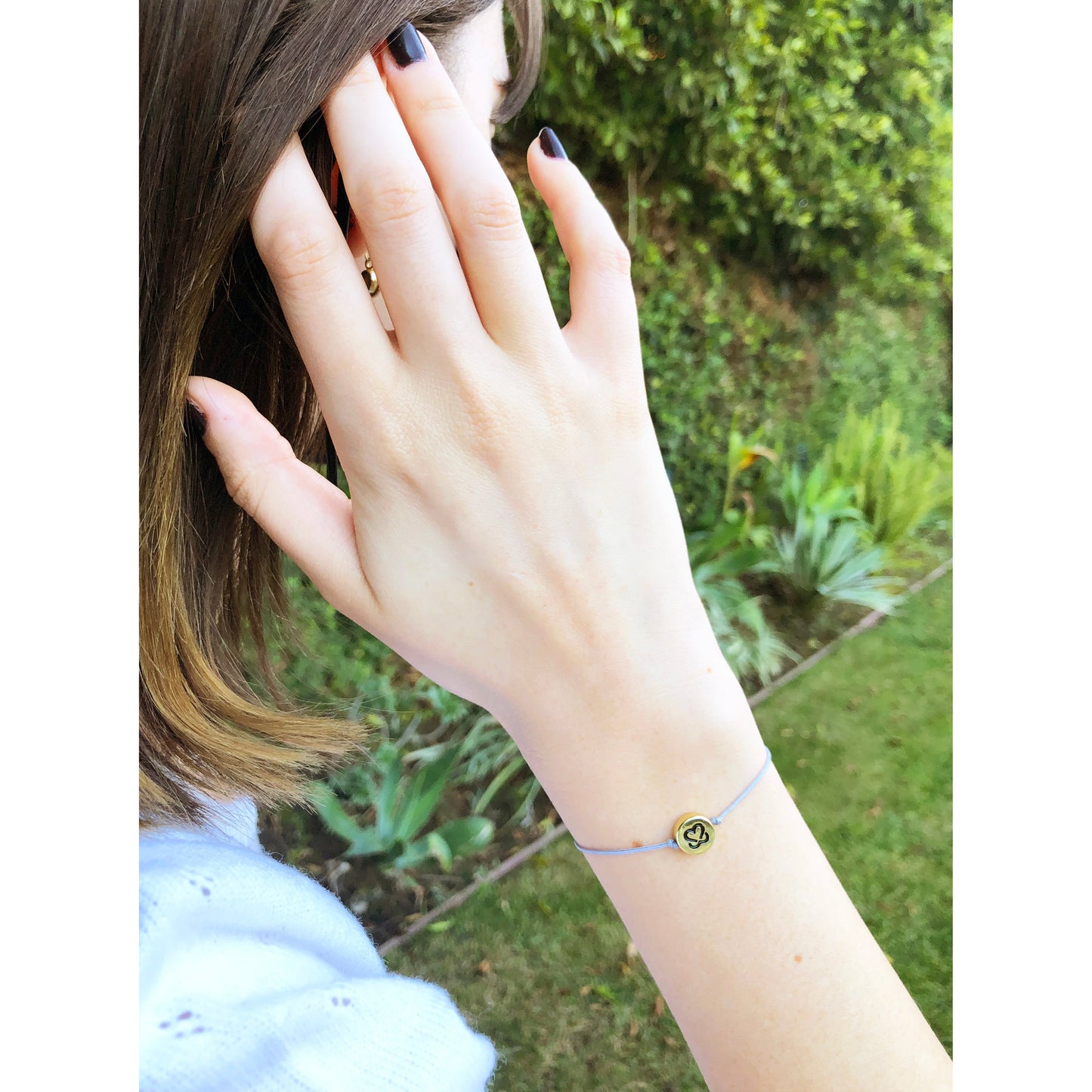 A woman's hand with dark nail polish rests near her face, showcasing a delicate blue string bracelet with a small gold charm.